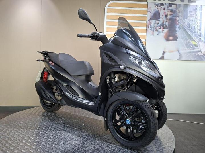 A sleek black three-wheeled scooter displayed on a round platform, with modern design features and a city-themed background.