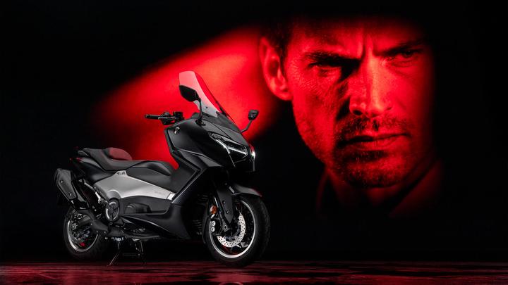 A sleek black and silver motorcycle is displayed prominently in the foreground, with a dramatic red-lit portrait of a man’s intense expression in the background. The setting has a dark ambiance, enhancing the motorcycle's features and the emotional impact of the portrait.