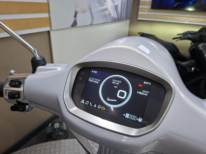 A close-up view of a scooter's dashboard displaying a digital screen with information including average consumption of 72.2 mpg, a range of 37 miles, current temperature of 24°C, and an odometer reading of 1750 miles. The dashboard is located on a light-colored scooter, with buttons and controls visible on the left side.