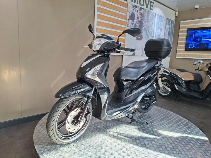 A black scooter displayed on a circular platform inside a showroom, with a light-colored wall in the background and another scooter partially visible on the right.