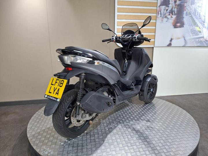 A black three-wheeled scooter displayed on a circular platform, with a registration plate visible. The background features a neutral wall and an image of a busy street.