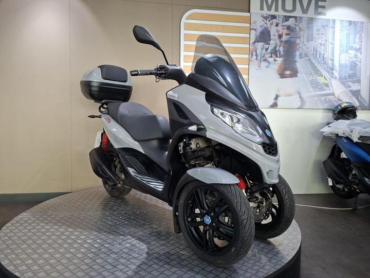 A gray and black three-wheeled scooter displayed on a rotating stand indoors, with a rear storage compartment and a large windshield. The background features an urban-themed wall with a move-related sign.
