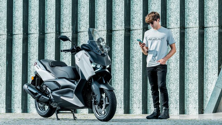 A young man in a gray t-shirt and black jeans stands beside a modern silver scooter, looking at his smartphone. The background features textured, vertical panels.