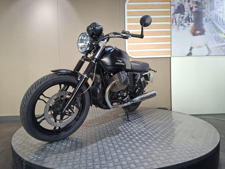 A black motorcycle is displayed on a round metal platform, showcasing its sleek design with a prominent headlight, chunky tires, and visible engine components, in front of a neutral-colored backdrop.