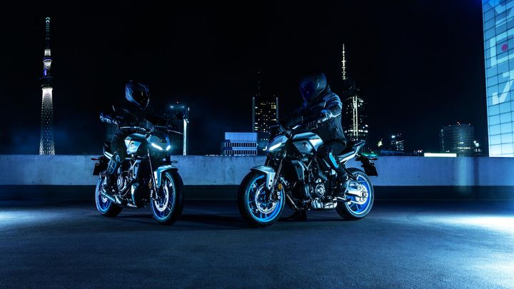 Two stylish motorcycles with illuminated features are parked on a rooftop at night, with a skyline featuring tall buildings and a tower in the background.