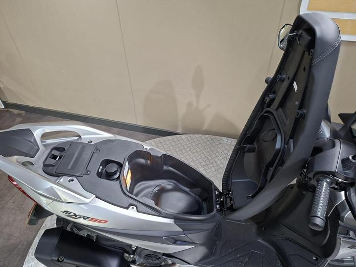 A close-up view of the seat compartment of an SRX 50 scooter, showing the area beneath the seat with storage options and a partially open lid. The background features a light-colored wall.