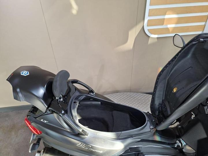 A close-up view of the storage compartment of a motorcycle with the seat slightly open, revealing a black interior and a sleek, dark exterior. The motorcycle features a logo and a shiny finish, set against a neutral backdrop.