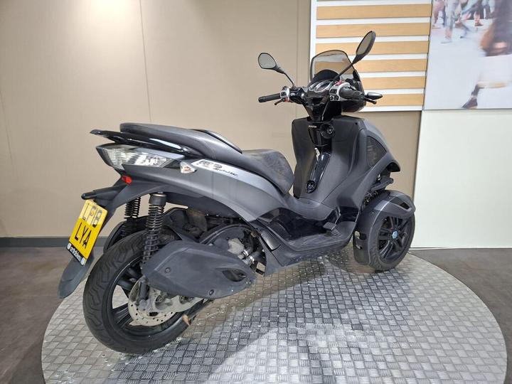 A black three-wheeled scooter parked on a textured circular platform, with a simple background featuring a wall and decorative wooden slats. The scooter has a sleek design, prominent front lights, and a license plate visible.