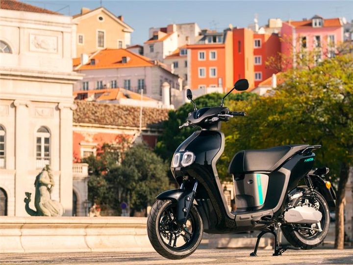 A sleek black electric scooter is parked in a lively urban setting, with colorful buildings and greenery in the background. The scene captures the essence of modern city life.