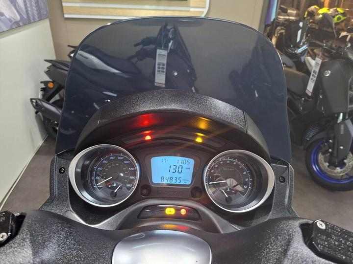 Close-up view of a scooter's dashboard displaying a speedometer, odometer showing 48,935 miles, and digital display with current speed of 130 mph. A dark tinted windshield is visible, along with warning lights indicating various statuses. Background features additional scooters.