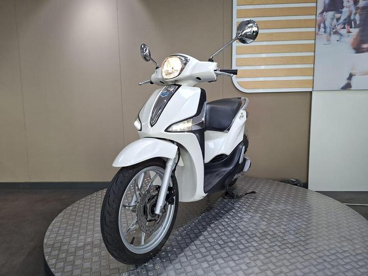 A white scooter displayed on a circular platform, featuring a sleek design, chrome mirrors, and a modern headlight emblem, set against a neutral background.