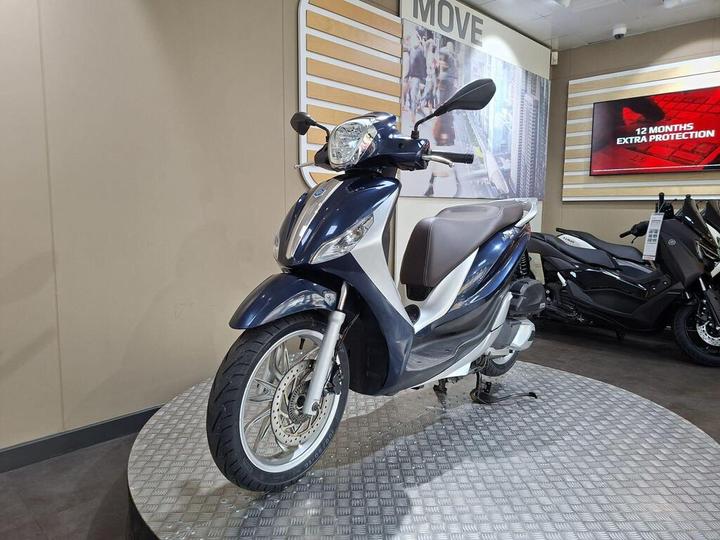 A blue and white scooter displayed on a circular metallic platform with promotional backgrounds in a showroom.