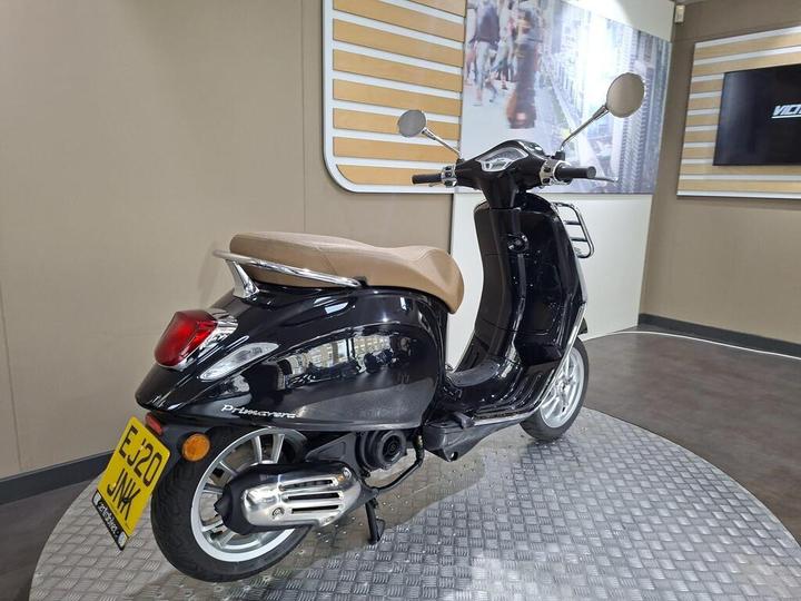 A black scooter with a tan seat, featuring a shiny rear end and chrome accents, positioned on a textured circular platform in a modern showroom setting.