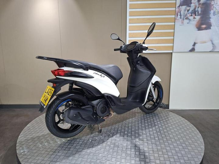 A white scooter with a sleek design, parked on a round metallic display. The scooter features a low profile, sporty rear design, and is positioned against a neutral wall backdrop.