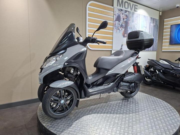 A gray three-wheeled scooter displayed on a circular platform in a showroom, featuring a front shield, a storage box on the back, and modern design elements. Other scooters are visible in the background.
