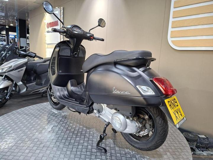 A black Vespa scooter is displayed on a circular metallic platform in a showroom, with a Yamaha sign visible in the background. Another scooter can be seen partially on the left side. The scooter features a sleek design with chrome accents and a rear license plate.