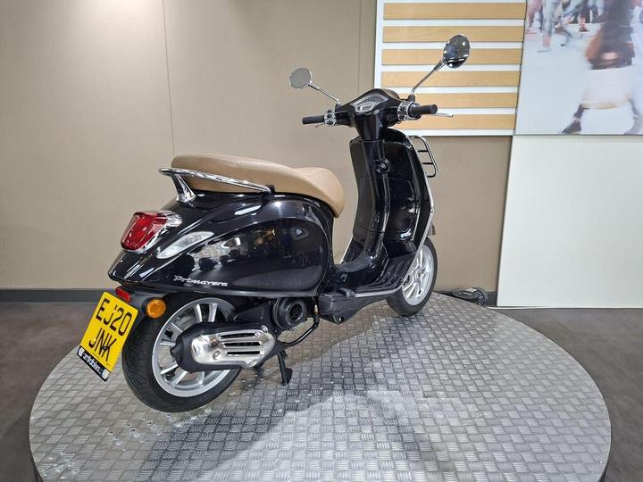 A black scooter with a tan seat is displayed on a circular platform, showcasing its rear and side profile. The license plate reads "EJ20 JNK." The background features a neutral wall and a blurred image of people.