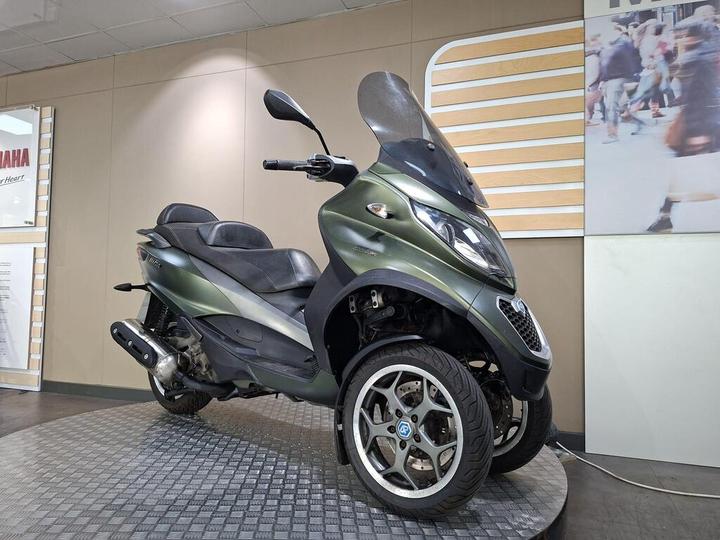 A green three-wheeled scooter displayed on a circular tiled platform, with a sleek design and a large windscreen, set against a light-colored wall and a blurred background of people.