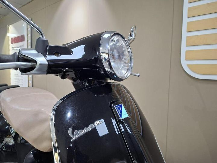 Close-up of a black Vespa scooter's front, showcasing the headlight, handlebars, and a seat in the background.