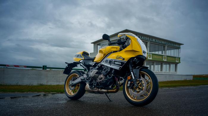 A yellow Yamaha motorcycle parked on a wet track, with a vintage-style pavilion in the background under a cloudy sky.