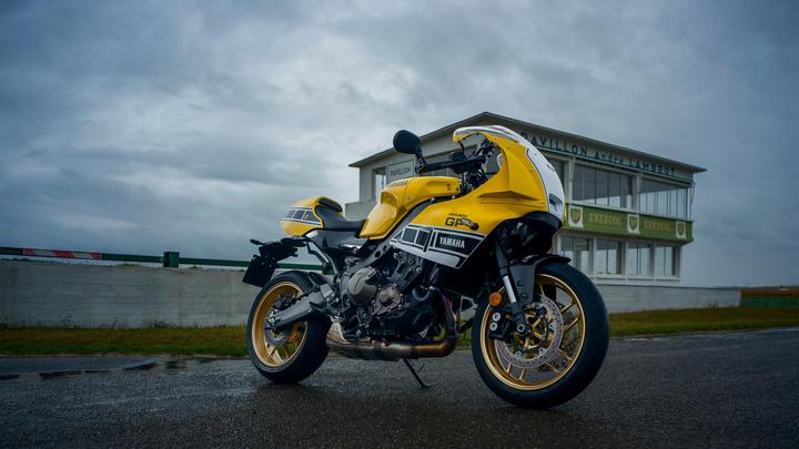 A yellow Yamaha motorcycle parked on a wet track, with a vintage-style pavilion in the background under a cloudy sky.