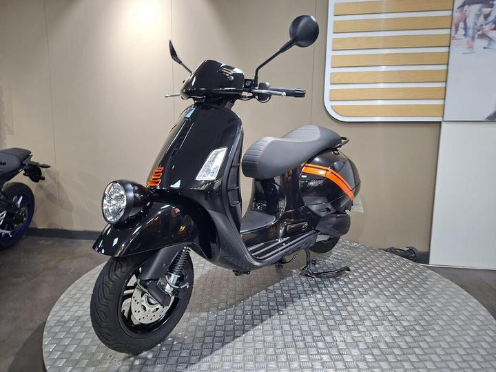 A sleek black scooter displayed on a circular platform, featuring stylish design elements such as an orange stripe on the rear and a comfortable gray seat. The background consists of a smooth, neutral-colored wall.