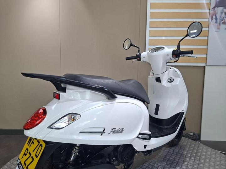 A white scooter with a sleek design is shown from the rear, highlighting its prominent rear light and dark seat. The handlebars feature mirrors, and the vehicle is positioned on a textured surface against a muted background.