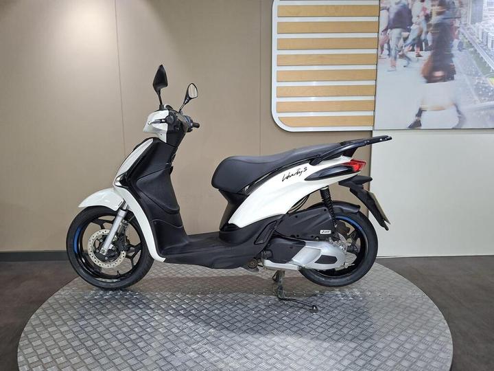 A white and black scooter displayed on a circular platform, featuring a modern design and prominent branding on the side. The background consists of a neutral wall with a partial image of people walking.