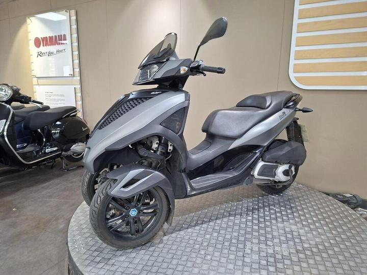 A gray scooter with three wheels parked on a display platform inside a showroom, with a Yamaha sign visible in the background.