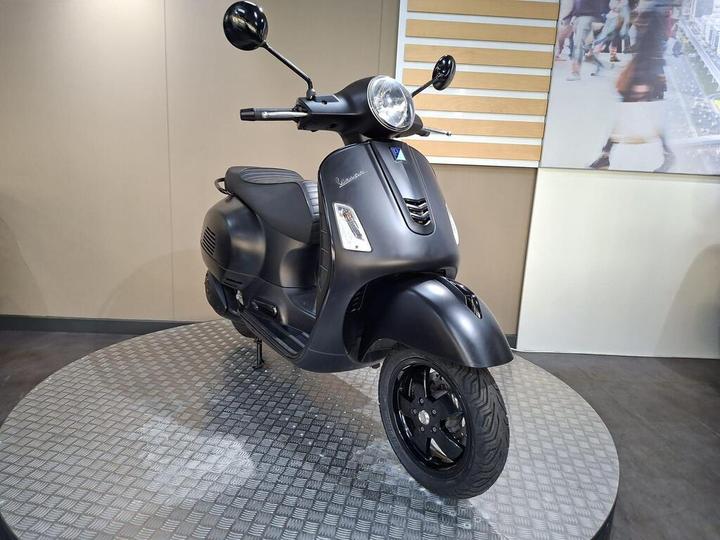 A black Vespa scooter displayed on a circular platform, with a sleek design and modern features, set against a neutral-colored wall.