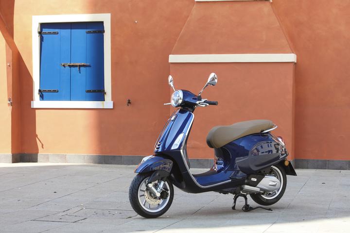 Left facing blue Vespa Primavera 50 with a orange wall and blue window backdrop